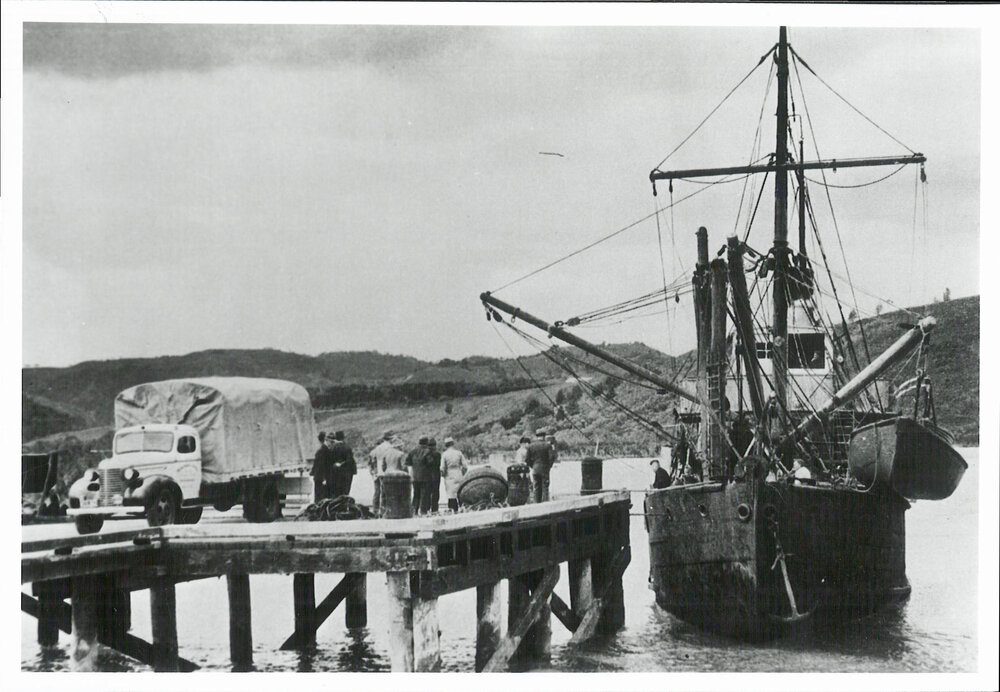 A group of men, dressed in coats and hats, stand near a large rugged wooden dock, a covered truck, and a moored ship. The truck features visible text on its side, though the specific wording is not discernible. The ship, equipped with masts and rigging, has a smaller boat attached to its side, adding to the maritime setting. In the background, hills with sparse vegetation frame the scene, suggesting a remote rugged area by a body of water. It is a black and white photo