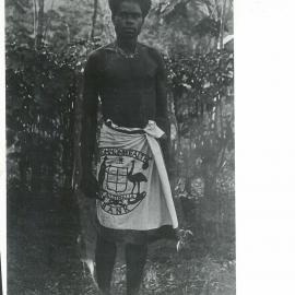 Demas - A member of the Rabaul Branch house staff, wearing the Commonwealth Badge suspended on a necklace of dogs' teeth. The “lava - lava” is an apron of white linen with the Bank's Coat of Arms worked in blue, c. 1920