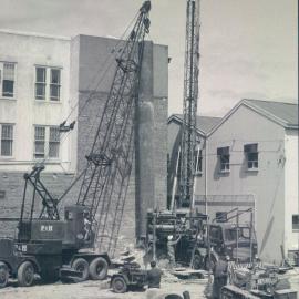 Adelaide Project - 1963-June 1967 - 3' liner being centred over shaft located alongside wall of A.W.U. building.  Drilling rig can be seen in background and a drilling "bit" in the foreground - 6 December 1963